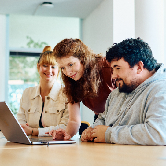 Eine Frau steht zwischen zwei sitzenden Mitarbeitenden und zeigt diesen etwas am Laptop, welcher auf dem Tisch vor ihnen steht.