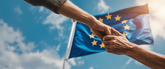 Zwei sich reichende Hände im Hintergrund weht eine Flagge der EU