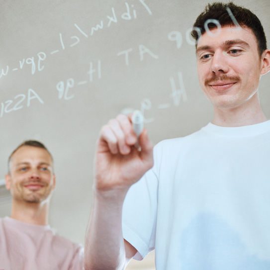 Zwei Mitarbeitende stehen vor einer Glasscheibe auf die sie mit einem weißen Stift Code darauf schreiben