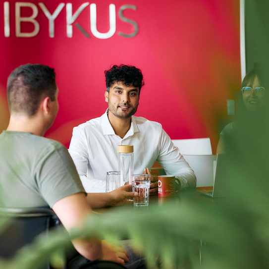 Zwei Mitarbeitende sitzen gemeinsam in der Cafeteria am Tisch, ein Mitarbeiter blickt dabei direkt in die Kamera. Im Hintergrund sieht man das IBYKUS Logo an einer roten Wand hängen.