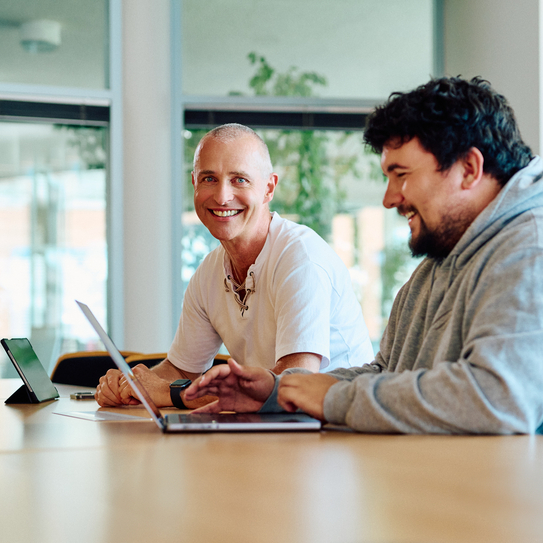 Zwei Mitarbeitende sitzen nebeneinander an einem Tisch, vor ihnen steht ein Laptop, die Person im Hintergrund schaut lächelnd in die Kamera