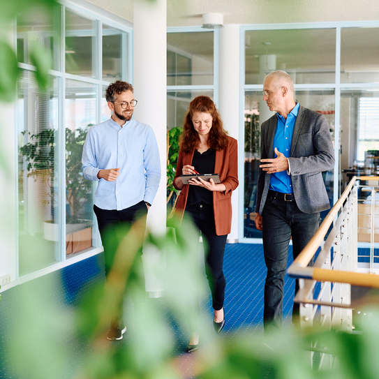 Drei Mitarbeitende laufen gemeinsam im Flur der IBYKUS AG auf einem blauen Teppich, dabei hält die Frau in der Mitte ein Tablet in der Hand.