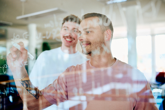 Zwei Mitarbeitende schreiben Code auf eine Glaswand