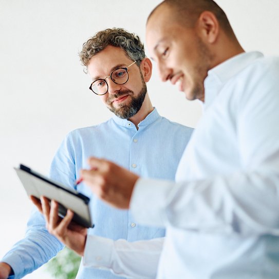 Zwei Mitarbeitende betrachten gemeinsam Inhalte auf einem Tablet, das der vorne stehende Mitarbeiter in der Hand hält.