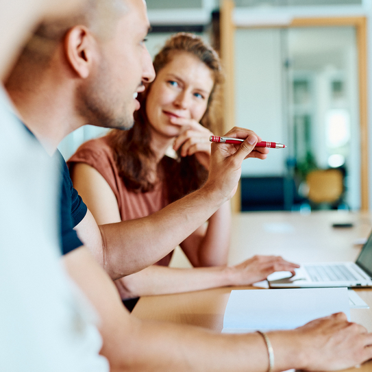 Drei Mitarbeitende sitzen an einem Tisch, im Hintergrund schaut eine Mitarbeiterin den Mitarbeitenden neben sich interessiert an. Dieser hält beim sprechen einen Kugelschreiber in der Hand.