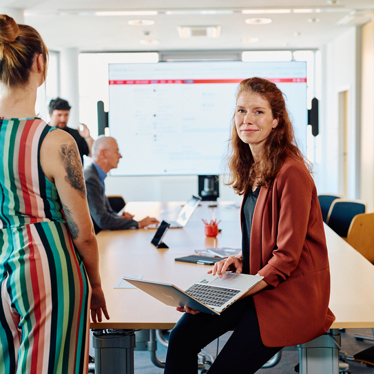 Eine junge Frau sitzt mit einem Laptop auf dem Schoß angelehnt auf einen Tisch, im Hintergrund sieht man andere Mitarbeitenden in einer Meetingsituation, auf dem Bildschimr im Hintergrund wird eine Präsentation gezeigt