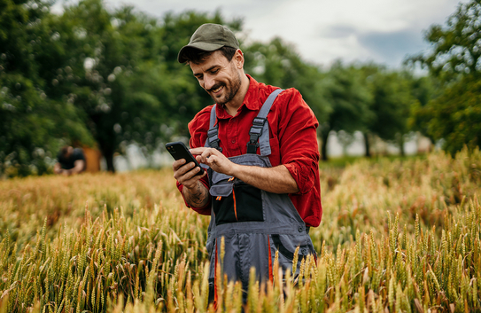 Ein Bauer steht mit seinem Handy in einem Feld, er trägt ein rotes Hemd und eine Latzhose