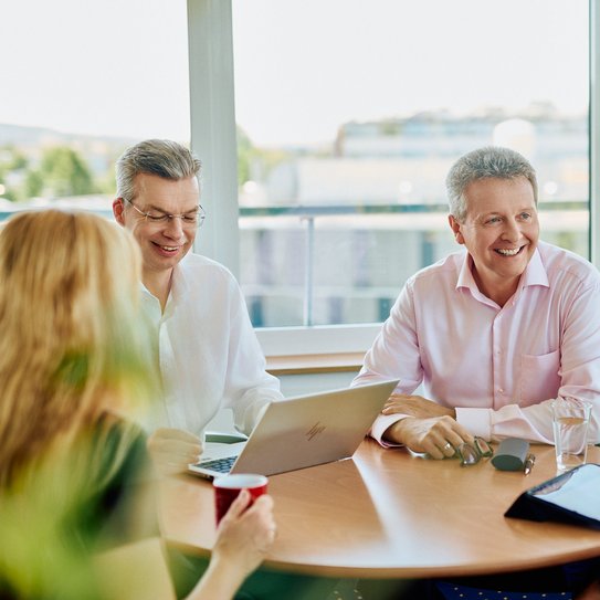 Drei Mitarbeitende sitzen in einem Meeting zusammen an einem runden Tisch.