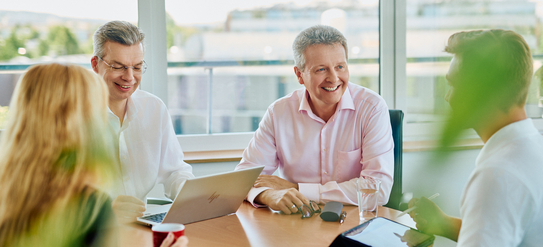 An einem Tisch sitze vier Personen, drei Männer und eine Frau sind in eine Unterhaltung vertieft. Auf dem Tisch steht ein aufgeklappter Laptop.