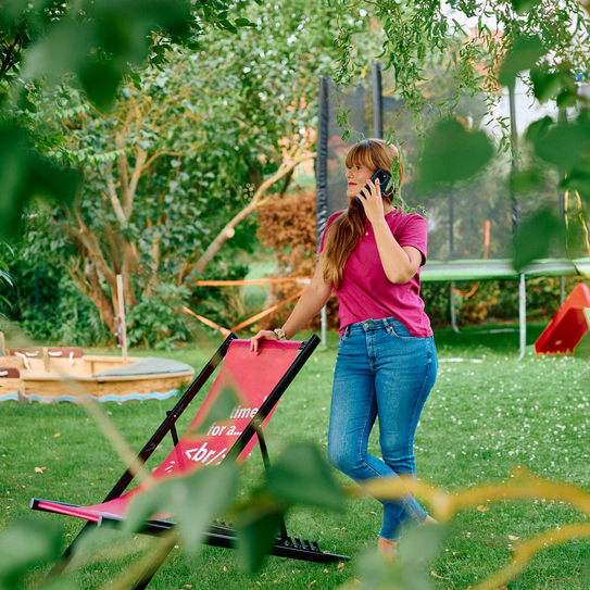 Frau telefoniert im Garten und lehnt sich an einen Liegestuhl.