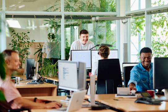 Mitarbeitende im Büro unterhalten sich über ihre Schreibtische hinweg