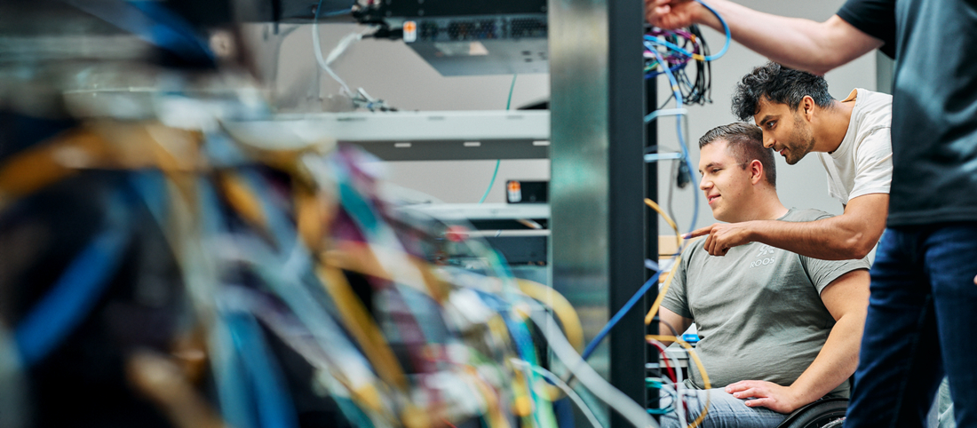 Ein Mitarbeiter zeigt einem Kollegen der im Rollstuhl sitzt, etwas an einem Servertower. Im Vordergrund sieht man angeschnitten einen anderen Mitarbeiter sowie einige Kabel.