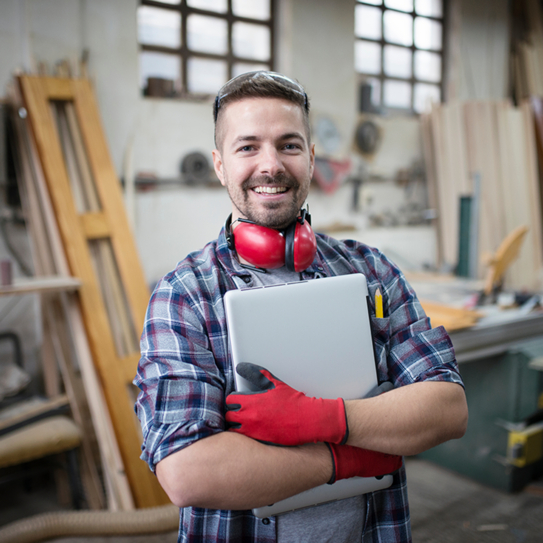 Ein Handwerker steht in seiner Werkstatt, er trägt einen gehörschutz um seinen ahls, in den Händen hält er einen Laptop