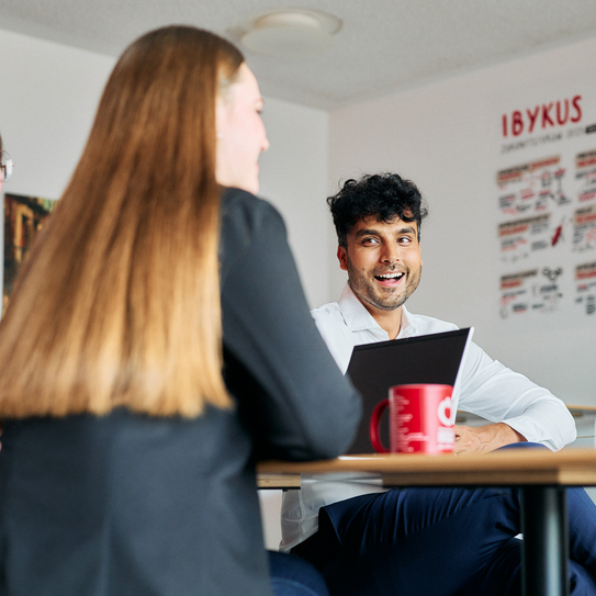 Eine Frau sitzt zusammen mit einem Kollegen an einem Tisch, sie unterhalten sich angeregt und lachend.