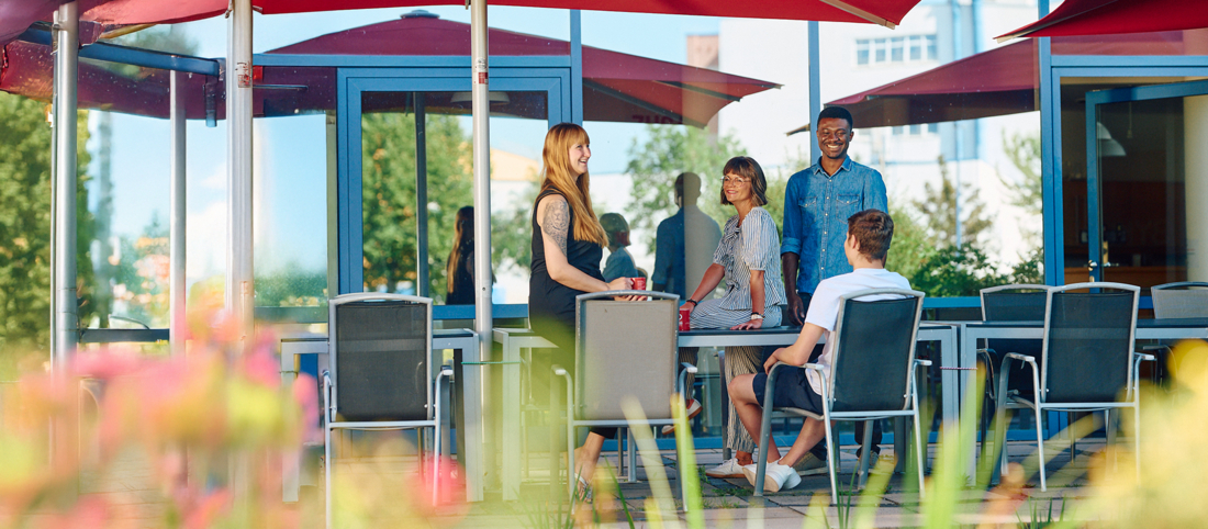 Diverse Menschen sitzen auf einer Terasse unter roten Sonnenschirmen. 