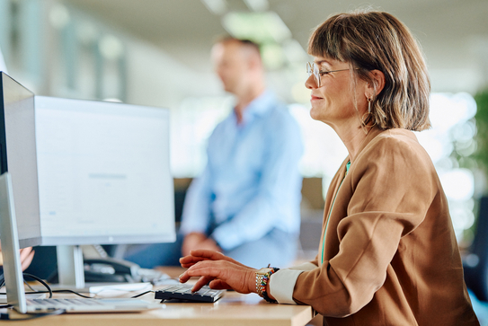 Eine Frau mit Brille schaut auf die beiden Bildschirme, welche vor ihr auf dem Schreibtisch stehen, dabei tippt sie etwas in die Tastatur