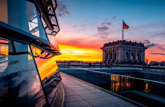 Ein Blick von der Glaskuppel des Bundestage in Berlin, im Hintergrund sieht man die Deutschlandfahne auf dem Gebäude. Der Himmel ist durch einen Sinnenuntergang gefärbt.