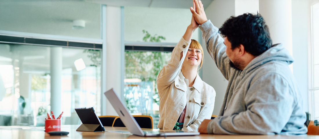 Zwei Mitarbeitende sitzen vor einem Schreibtisch und klatschen zum Highfive in die Hände