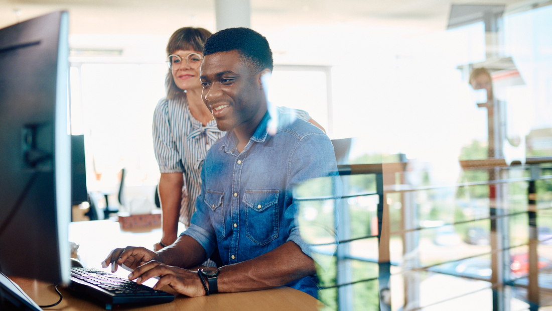 Ein Mann sitzt an einem Schreibtisch vor einem Bildschirm, er schaut dabei auf den Bildschirm. Neben ihm steht eine Kollegin die ebenfalls auf den Bildschirm schaut.