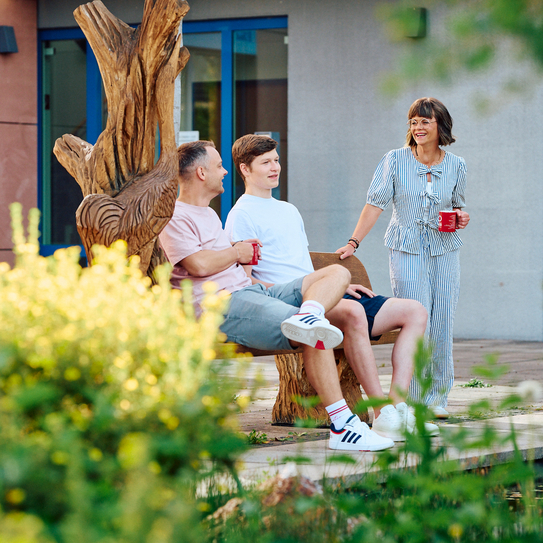 Zwei Mitarbeitende sitzen auf einer hölzernen Bank, daneben steht eine Mitarbeiterin mit einer Tasse in der Hand. Im Vordergrund sieht man Büsche und Pflanzen.