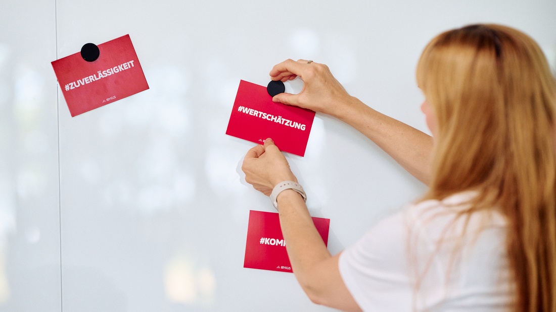 Eine Frau mit langen Haaren hängt eine Postkarte mit dem Wort Wertschätzung an einer Pinnwand auf
