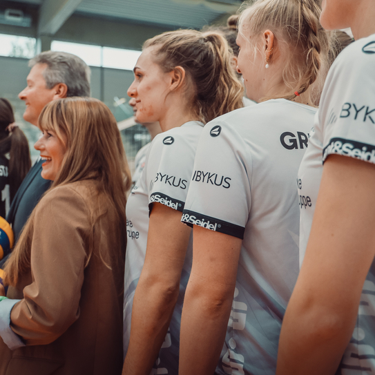 Ein paar Mitglieder des Volleyballteams stehen mit unserem Vorstand und unserer Marketingchefin zusammen, dabei tragen die Spielerin ihr Trikot mit IBYKUS Logo