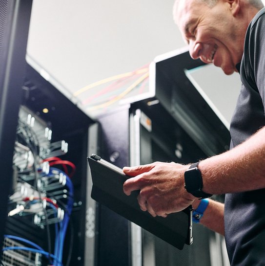 Techniker bei der Wartung eines Serverracks in einem Rechenzentrum – mit Tablet zur Systemüberwachung und modernen Netzwerkkomponenten im Hintergrund.