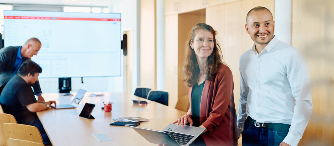 Ein Blick in einen Meetingraum, zwei Mitarbeitende sind im Vordergrund zu sehen. Sie lehnen an den Tisch an, an dem im Hintergrund noch zwei andere Mitarbeitende arbeiten.