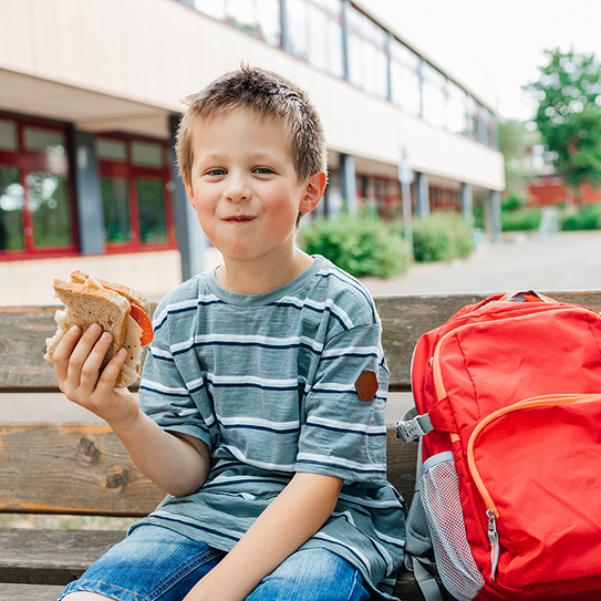 Ein kleiner Junge sitzt mit seinem roten Rucksack auf einer Bank und isst sein Schulbrot