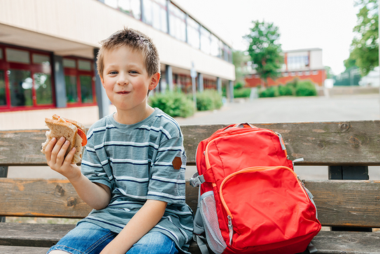 Ein Junge sitzt mit seinem Schulbrot auf einer Bank auf dem Schulgelände, neben ihm steht sein roter Rucksack