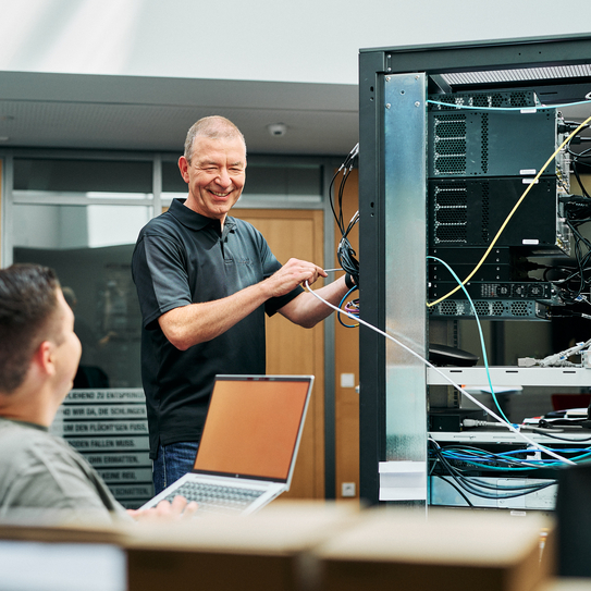 Eine Person steht vor einem Server Rack und lächelt einer anderen Person zu