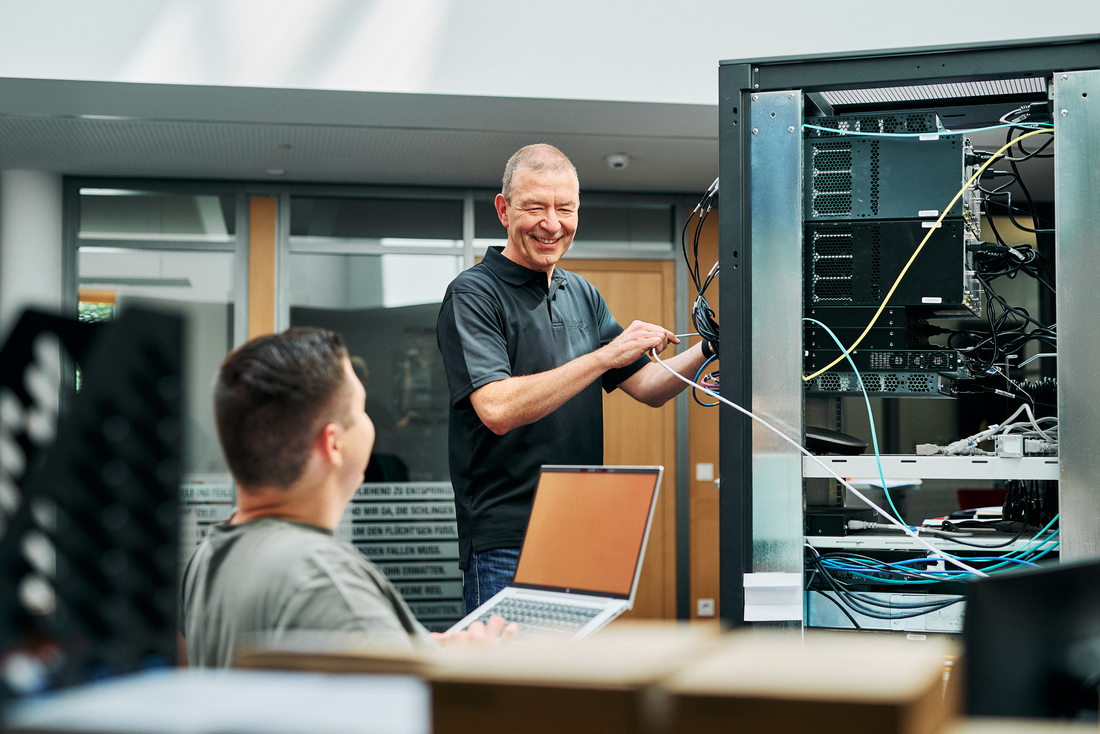 Zwei Mitarbeitende arbeiten gemeinsam an einem Serverturm, die Person am Server steckt mehrere Kabel an die Technik an